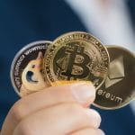 Mão segurando criptomoedas Bitcoin, Ethereum e Dogecoin