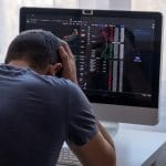 Homem jovem desesperado com as mãos na cabeça em frente ao computador com gráficos do bitcoin