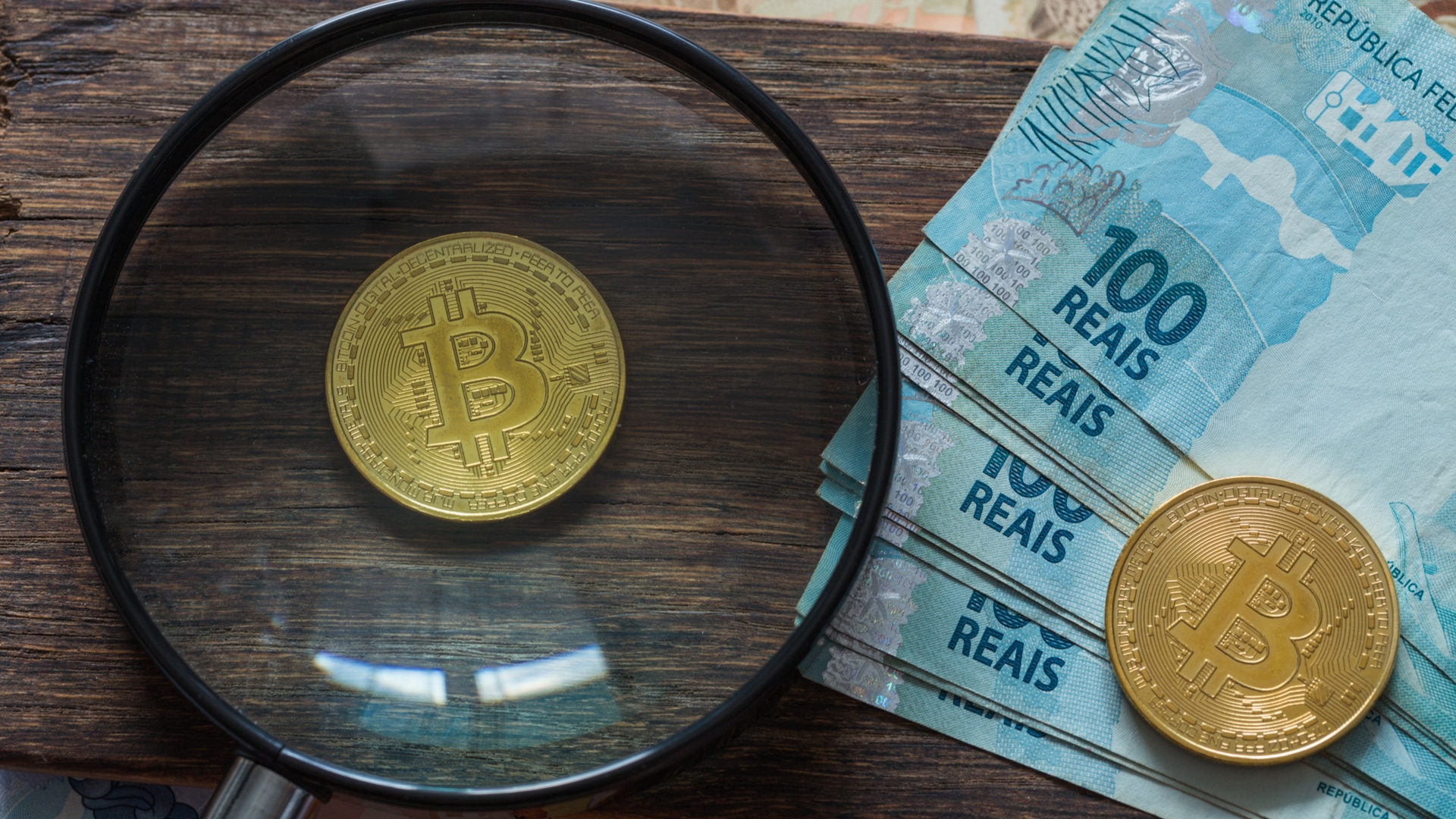 Lupa de olho em Bitcoin próxima de notas de Real do Brasil