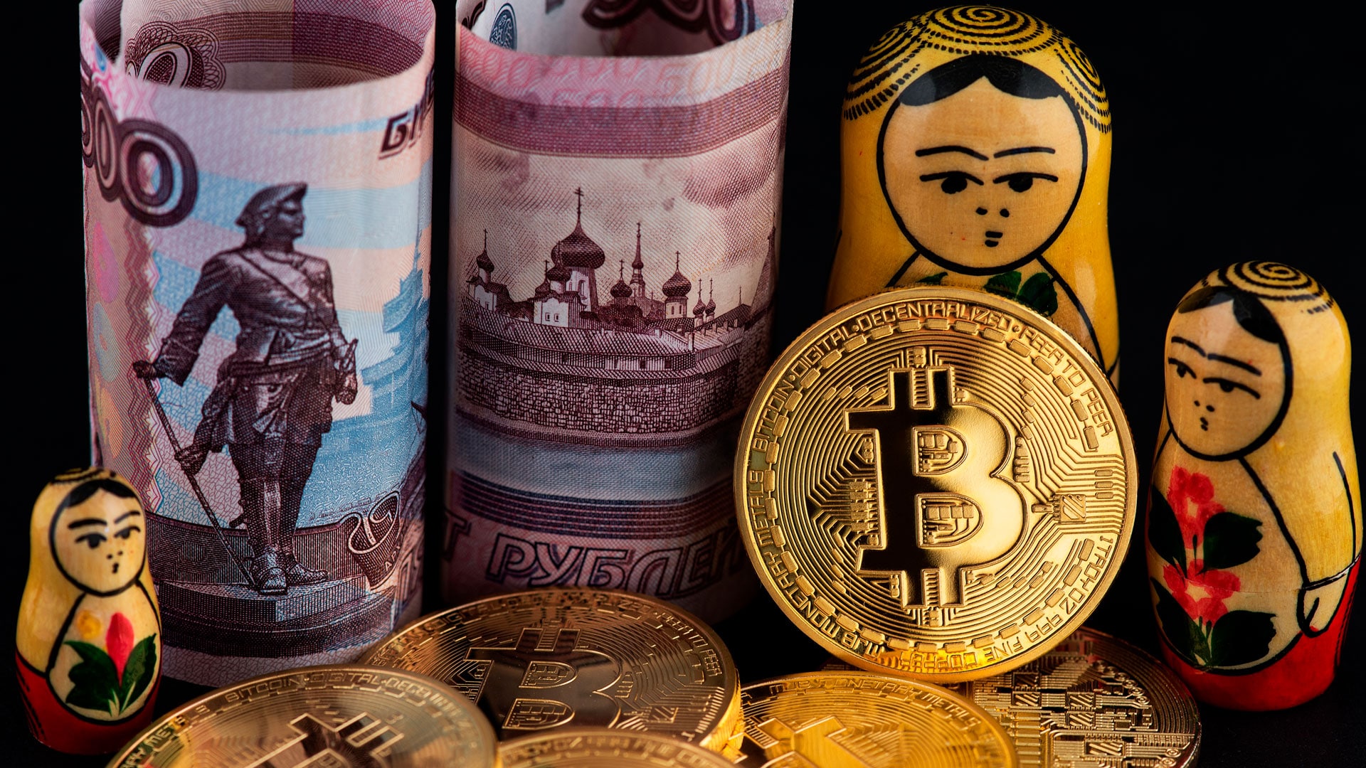 Moedas físicas de Bitcoin ao lado de rublos da Rússia e matrioskas.