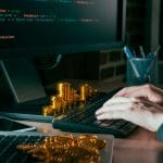 Hacker escrevendo código ao lado de pilha de criptomoedas.