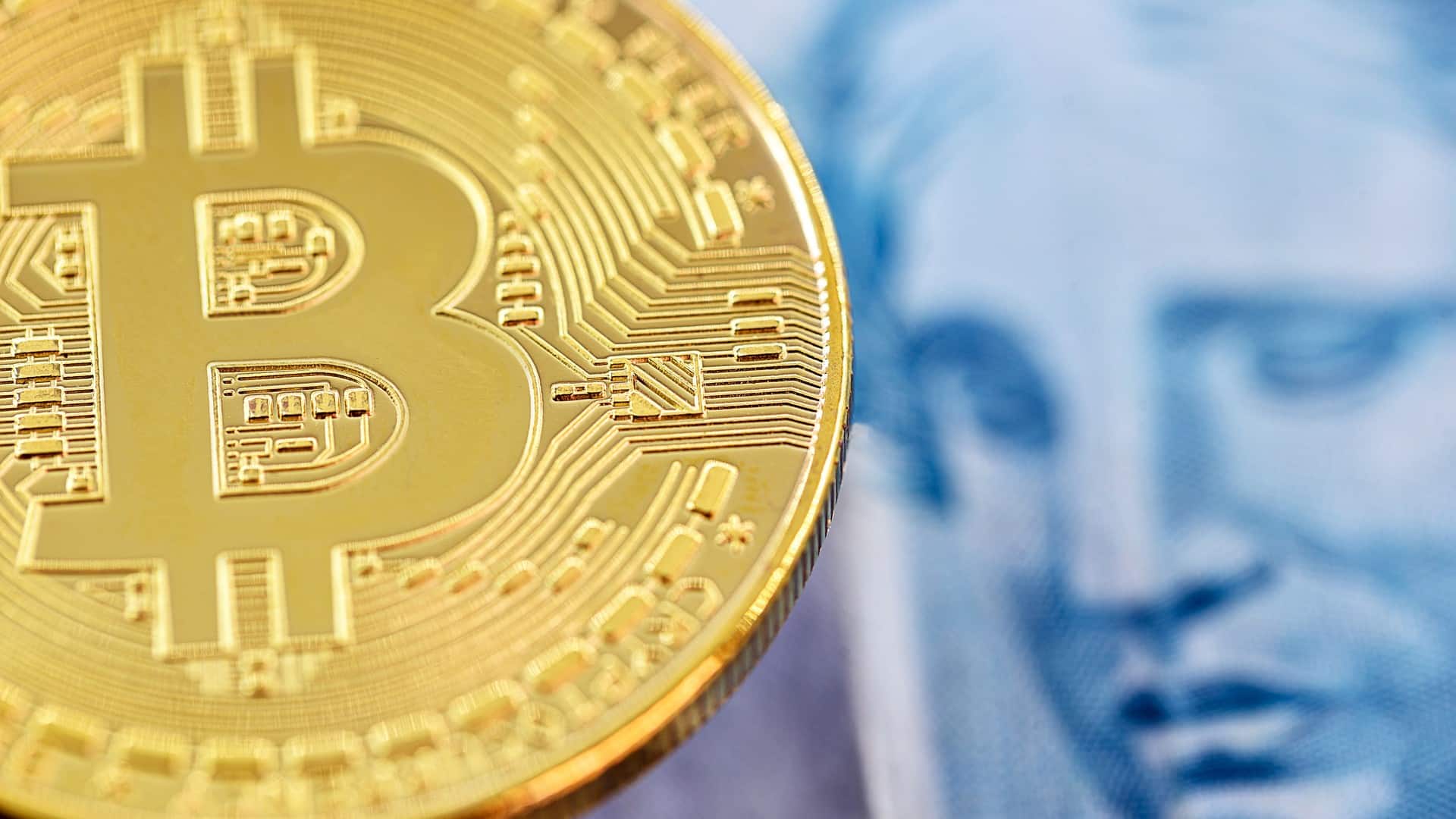 Bitcoin ao lado de nota de Real brasileiro, moeda emitida pelo Banco Central do Brasil