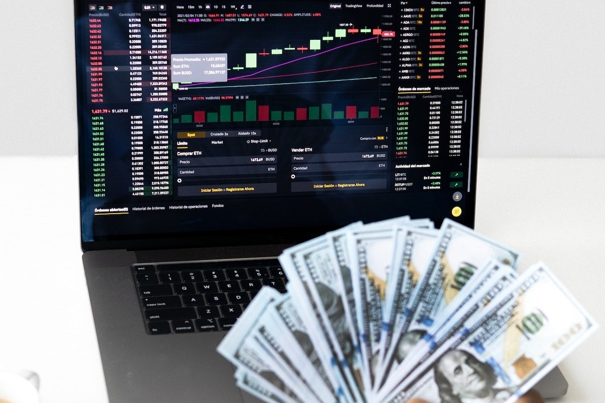 Homem segurando notas de dinheiro em frente a notebook com tela gráfica de preços de criptomoedas