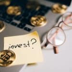 Bitcoin com papel ao lado perguntando sobre investimentos, em cima de um notebook