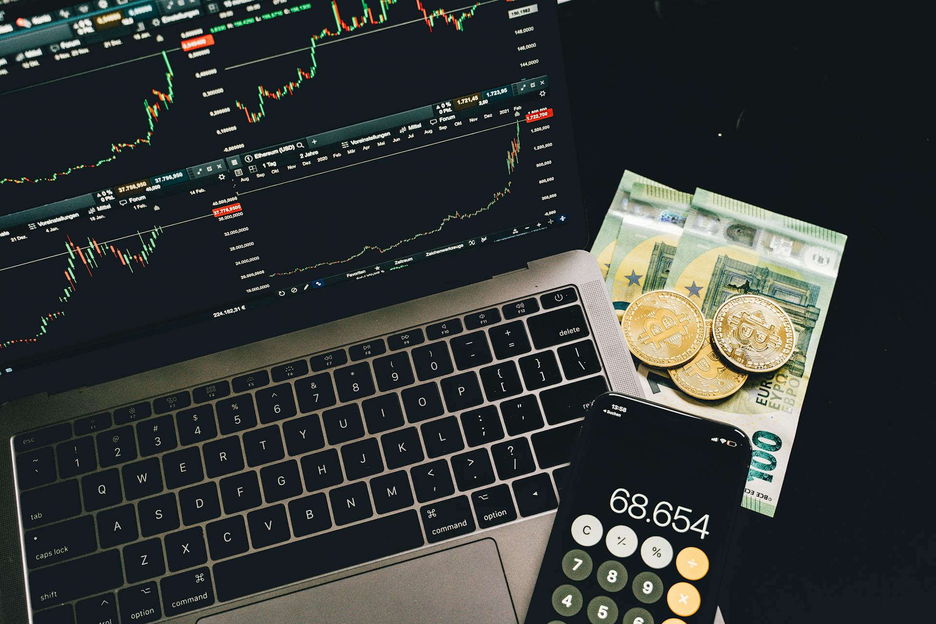 Moedas de bitcoin sobre notas de Euro, ao lado de computador em tela de negociações