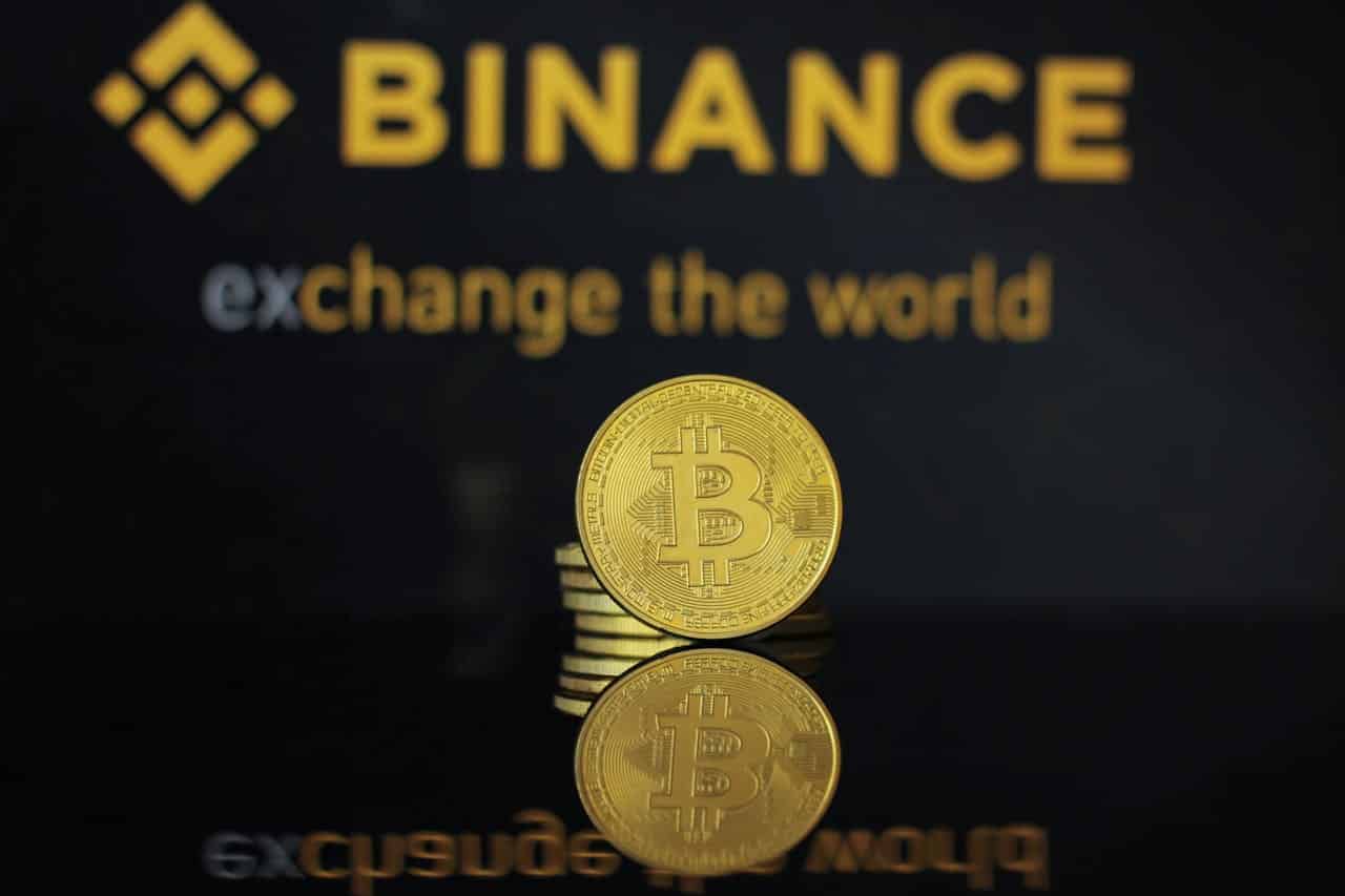 Logo da Binance ao fundo, com bitcoin simbólico a frente